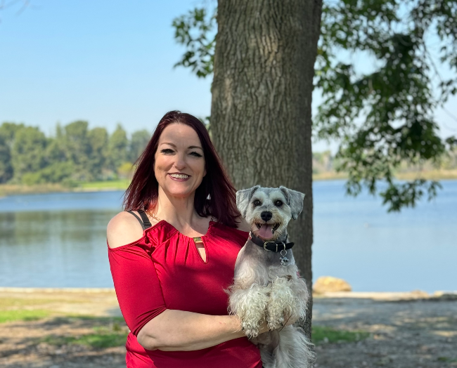 Valeri Gragg Dog Groomer holding a white dog next to a tree and body of water